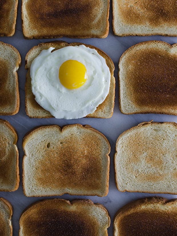 Várias fatias de torradas de pão de forma branco, e um ovo frito em cima de uma delas. vistos de cima, sobre uma mesa branca.